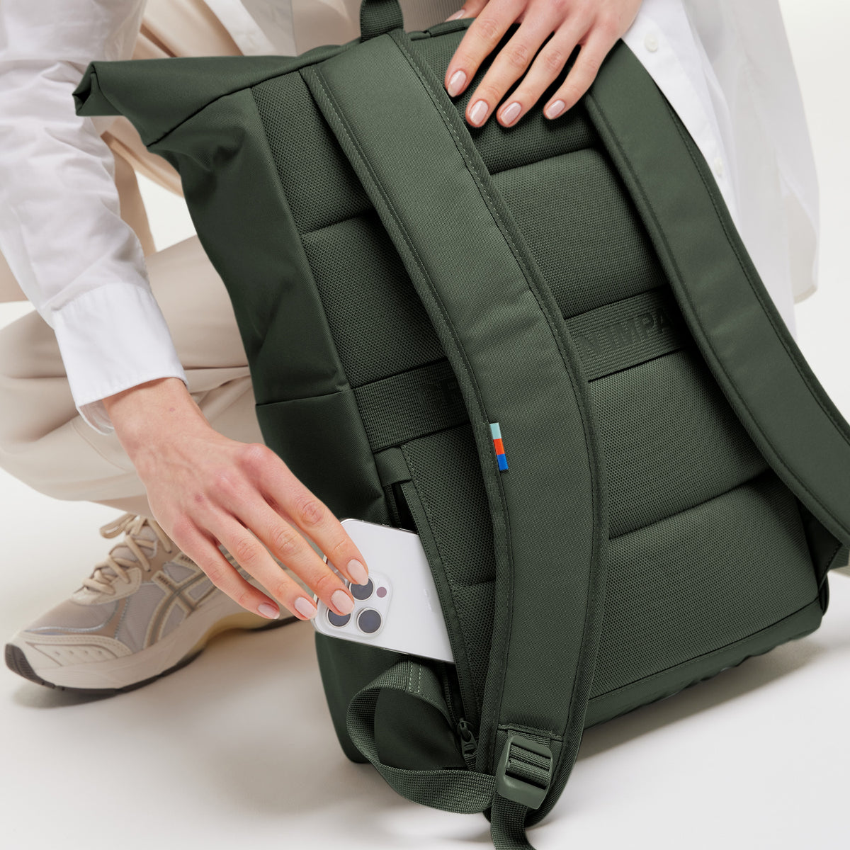 Eine Frau steckt ein Telefon in die versteckte Seitentasche des ROLLTOP EASY Rucksacks in algae (Dunkelgrün mit Graustich).