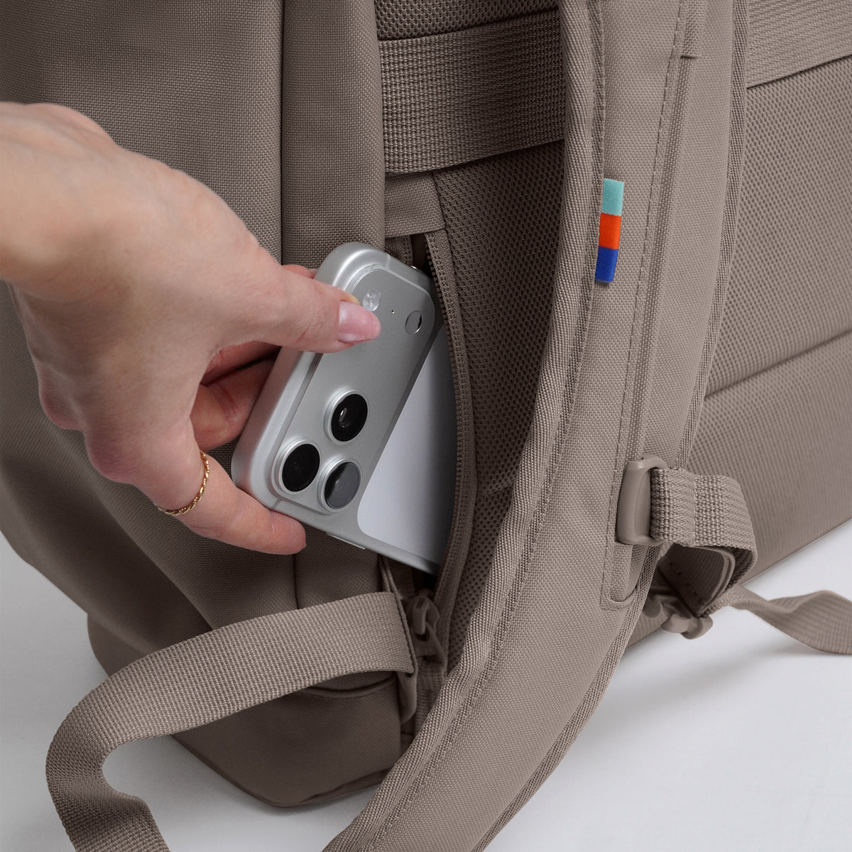 FLAP PACK LARGE Rucksack in oyster (klassisches Taupe-Braun) mit Smartphone, das von einer Frau in die Seitentasche gesteckt wird.