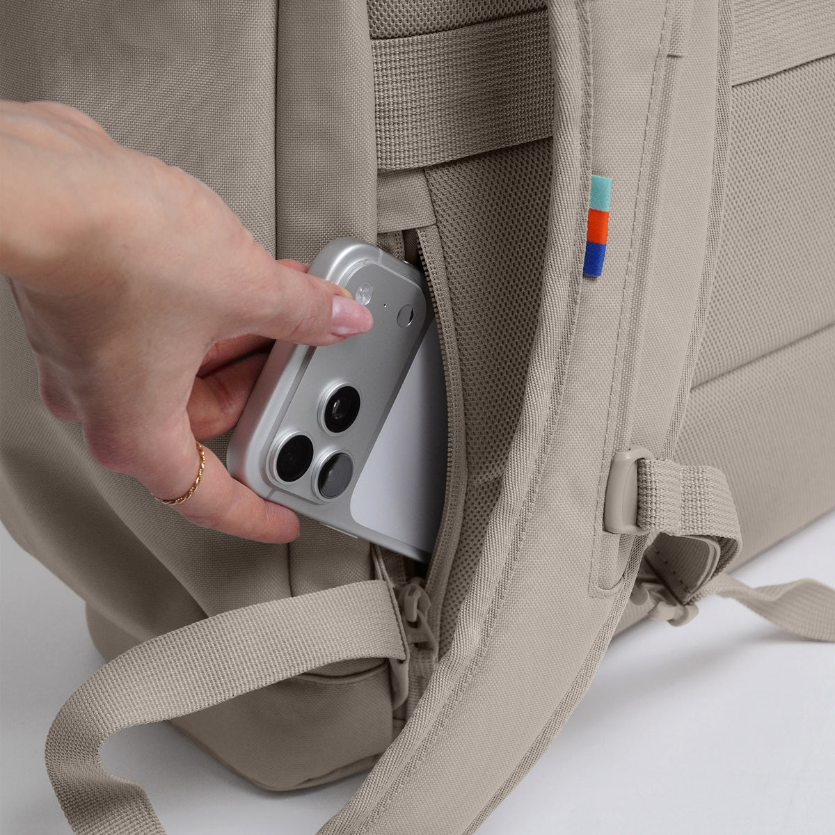 FLAP PACK LARGE Rucksack in scallop (helles Beige) mit Hand, die Smartphone in versteckte Seitentasche steckt.