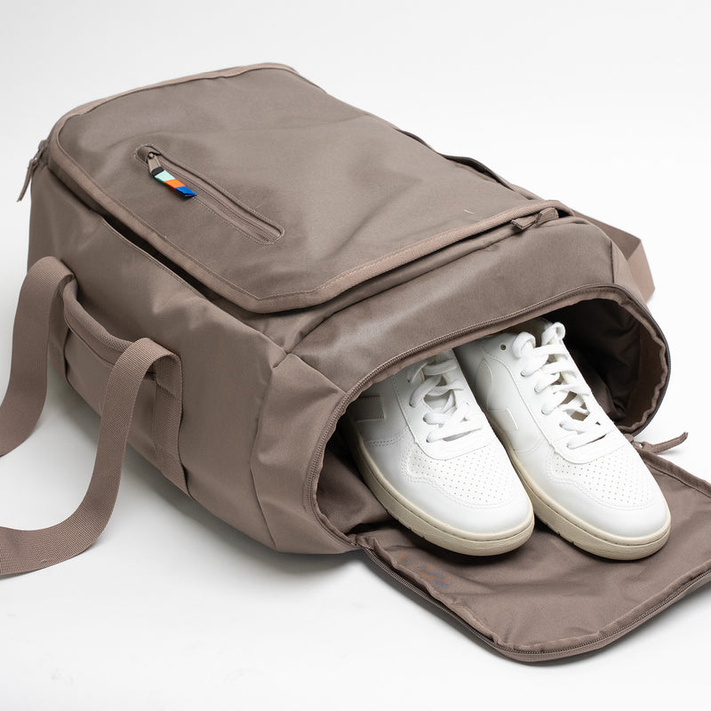 DUFFLE PACK Tasche in oyster (klassisches Taupe-Braun) mit weißen Sneakern im geöffneten Seitenfach.