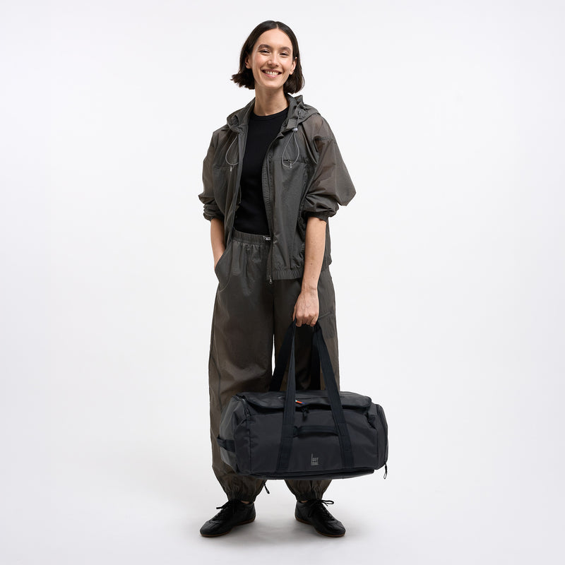 DUFFLE PACK Tasche in black (Schwarz) von einer Frau in der Hand gehalten.