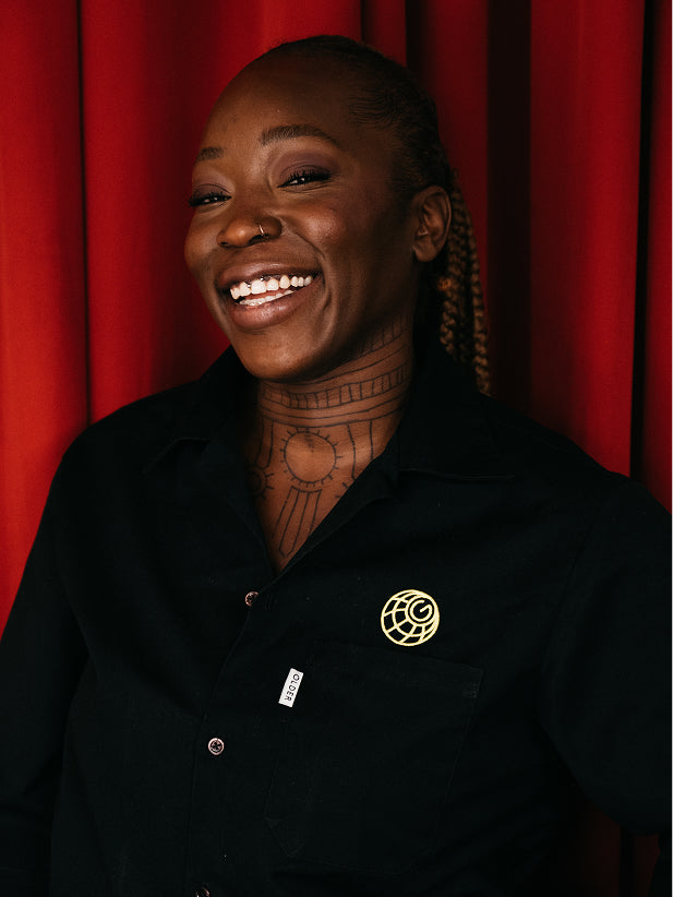 A golden, geometric accessory as a brooch on a black shirt, worn by a smiling woman.