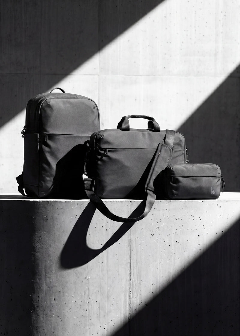 Tre accessori: zaino, borsa a tracolla e custodia su una superficie di cemento con forti ombre diagonali.