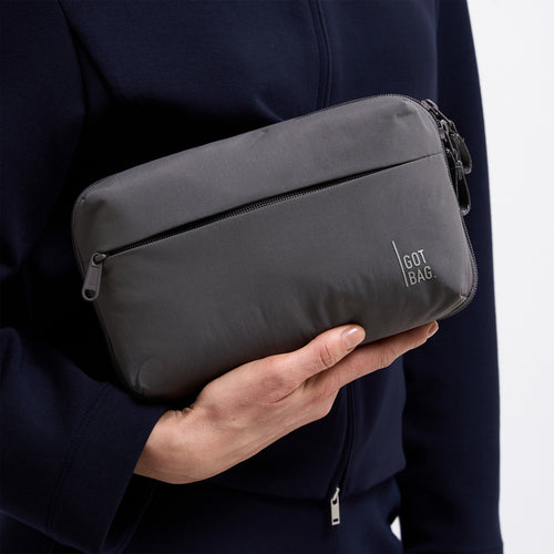 COMMUTER TECH POUCH Tasche in shark (Dunkelgrau) von einem Mann in der Hand gehalten.
