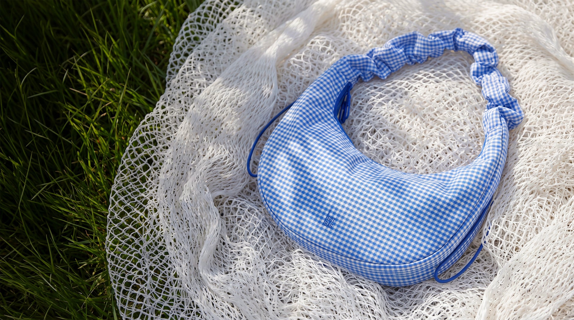 MOON BAG RUFFLE VICHY tas in vichy cobalt (Blauw-beige geruit) liggend op lichte netstof.