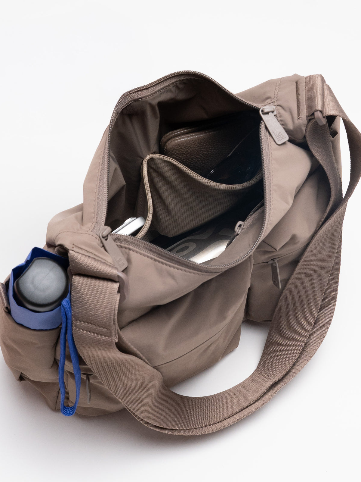 POCKET SLING BAG Tasche in oyster (klassisches Taupe-Braun) mit Einblick ins gefüllte Innere und Trinkflasche.