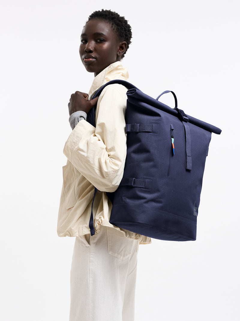 ROLLTOP Rucksack in tide (tiefes Dunkelblau mit Lilastich) von einer Frau auf dem Rücken getragen.