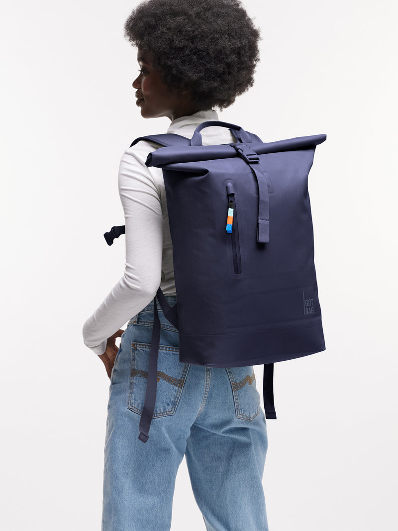 ROLLTOP LITE Rucksack in tide (tiefes Dunkelblau mit Lilastich) von einer Frau auf dem Rücken getragen.