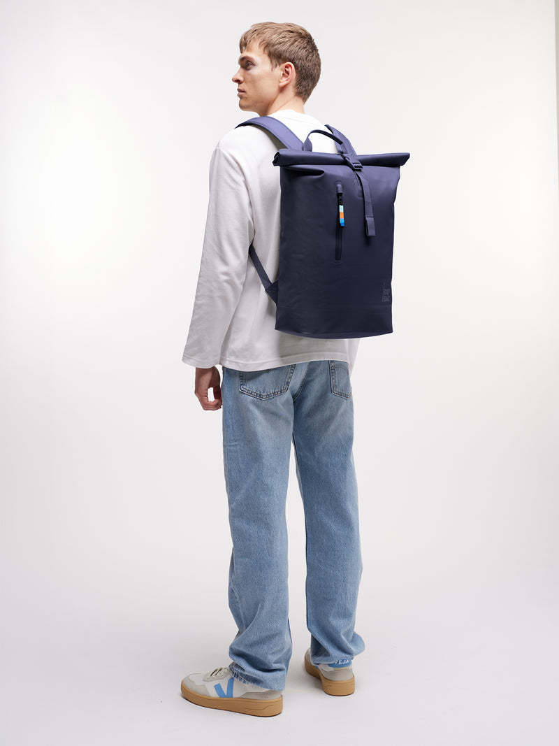 ROLLTOP LITE Rucksack in tide (tiefes Dunkelblau mit Lilastich) von einem Mann auf dem Rücken getragen.