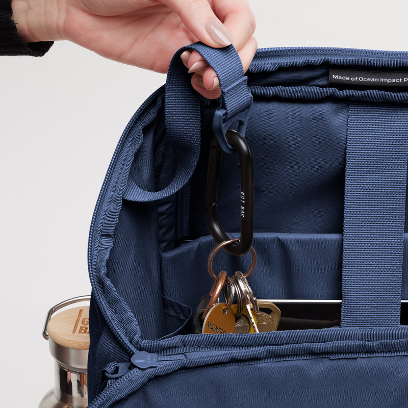 PRO PACK MONOCHROME Rucksack in deep ocean (dunkles Marineblau), von einer Frau am Gurt gehalten, mit Schlüsselbund.