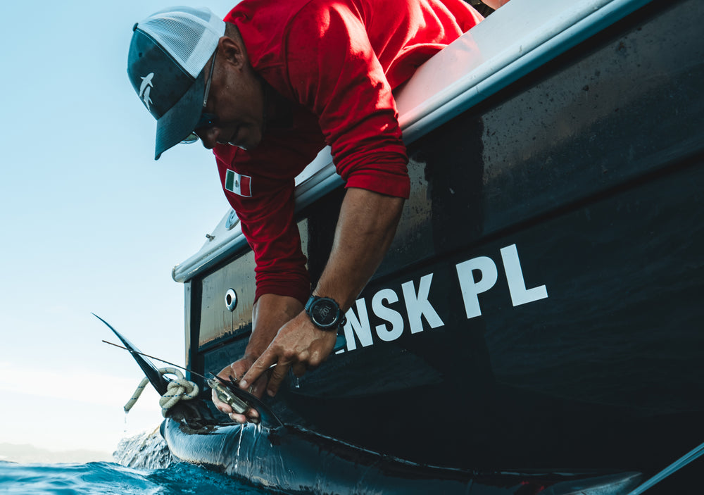 Uomo su una barca che indossa accessori (orologio, cappellino) e si prende cura di un pesce nell'acqua.