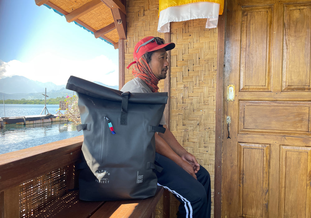 GOT BAG backpack next to a man on a wooden bench by the sea, with mountains in the background.