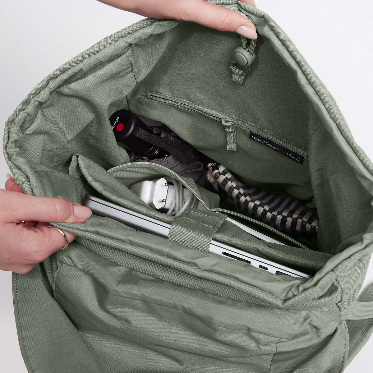 FLAP PACK Rucksack in bass (Salbeigrün), von Händen geöffnet, zeigt Innenleben mit Laptop und Regenschirm.