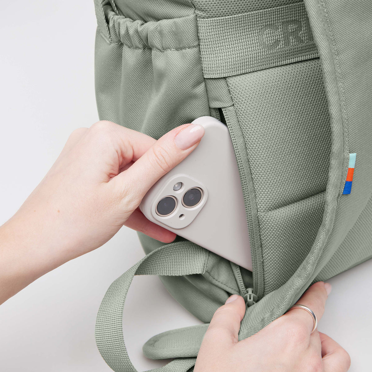 FLAP PACK Rucksack in bass (Salbeigrün). Eine Frau steckt ein Handy in die Seitentasche.