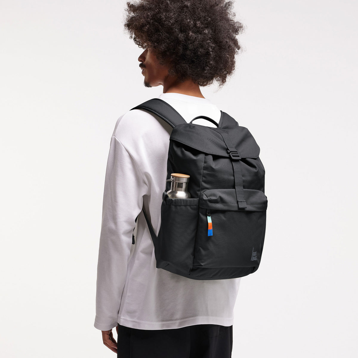 FLAP PACK Rucksack in black (Schwarz) von einem Mann auf dem Rücken getragen, mit Wasserflasche in der Seitentasche.