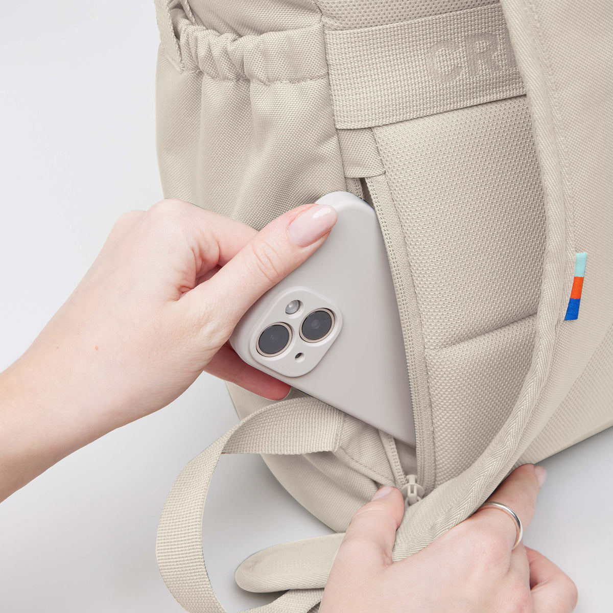 FLAP PACK Rucksack in soft shell (warmes Cremeweiß), von einer Frau ein Smartphone in die Reißverschlusstasche gesteckt.