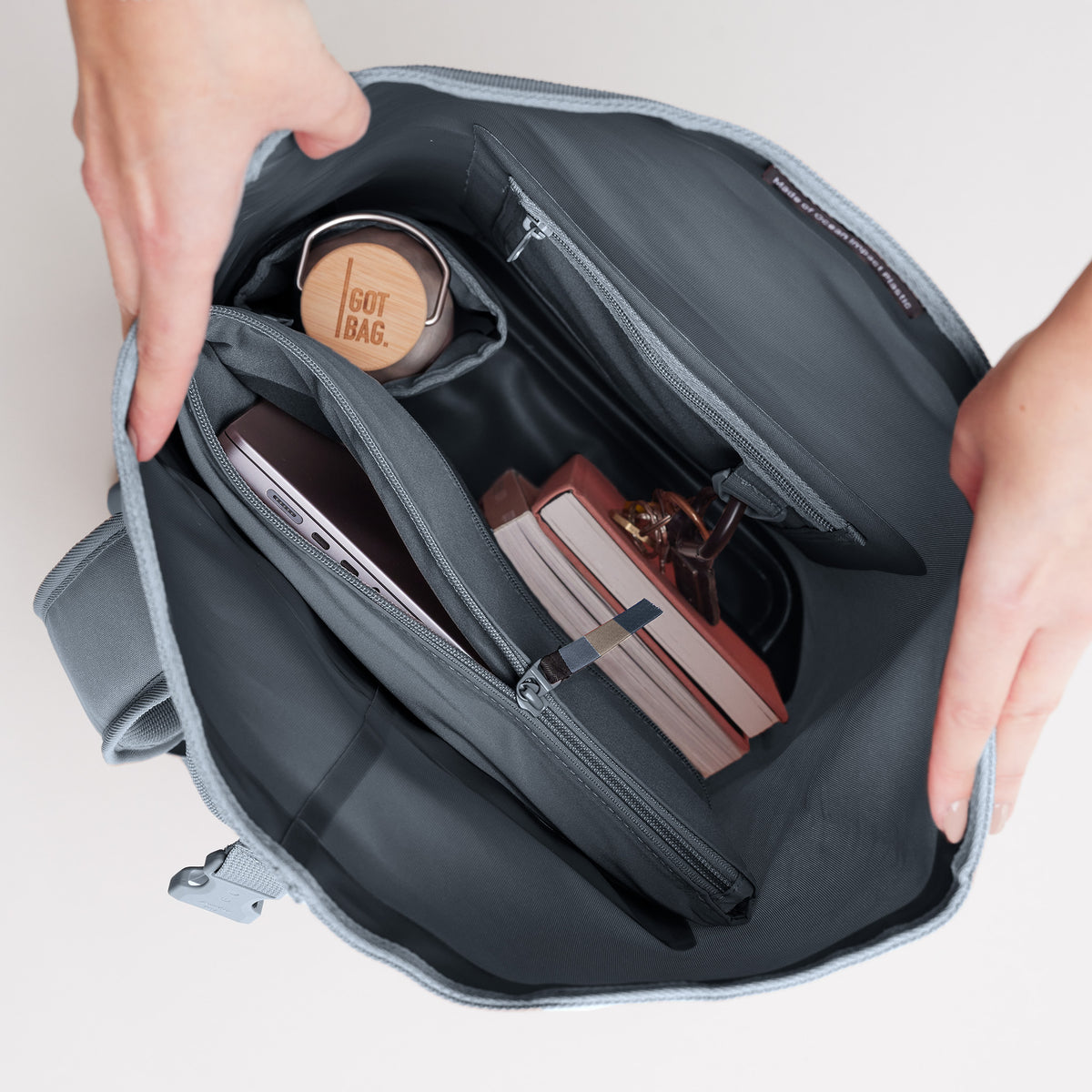 ROLLTOP LITE Rucksack in marlin (grau-blau) von Händen geöffnet, Innentaschen mit Laptop, Trinkflasche und Büchern.