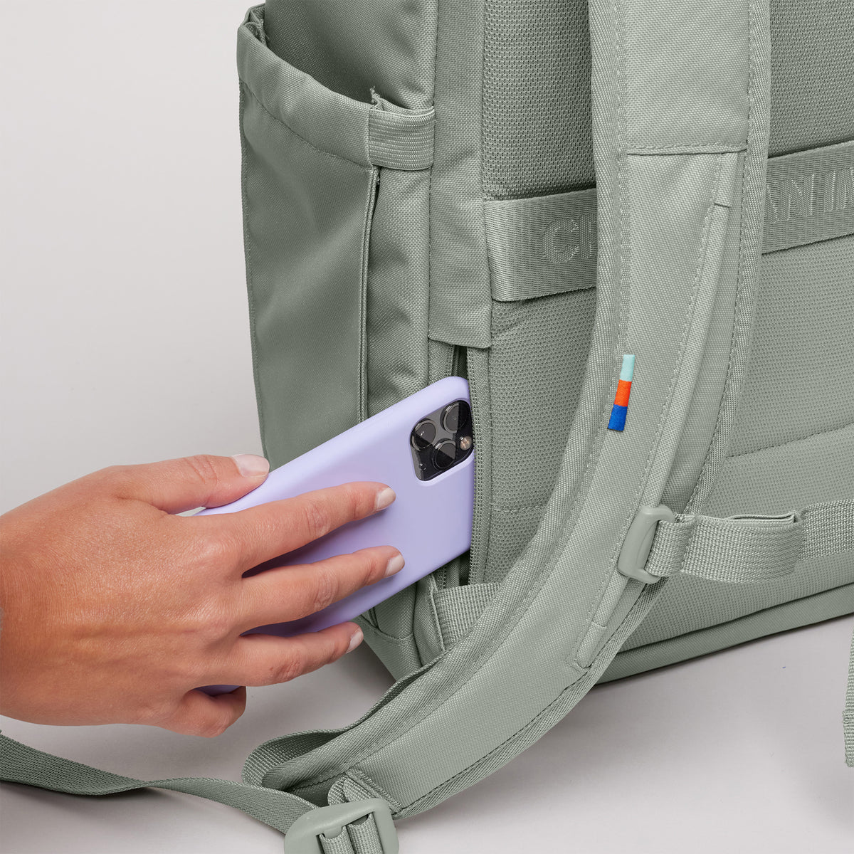 SERENE PACK Rucksack in bass (Salbeigrün), eine Hand steckt ein lila Telefon in eine Seitentasche.