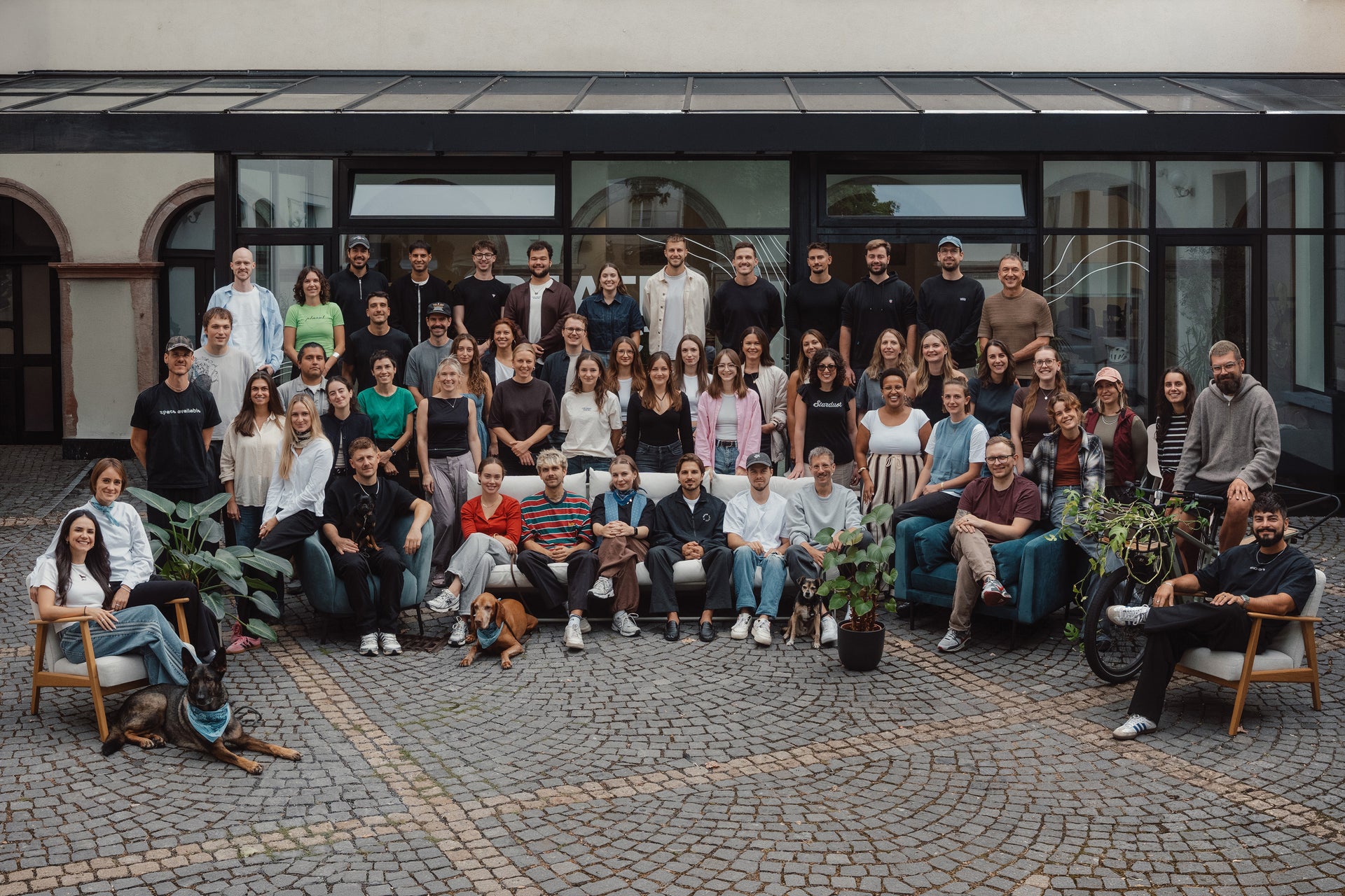 Grote teamfoto van mannen en vrouwen die buiten poseren op een geplaveide binnenplaats voor een gebouw.