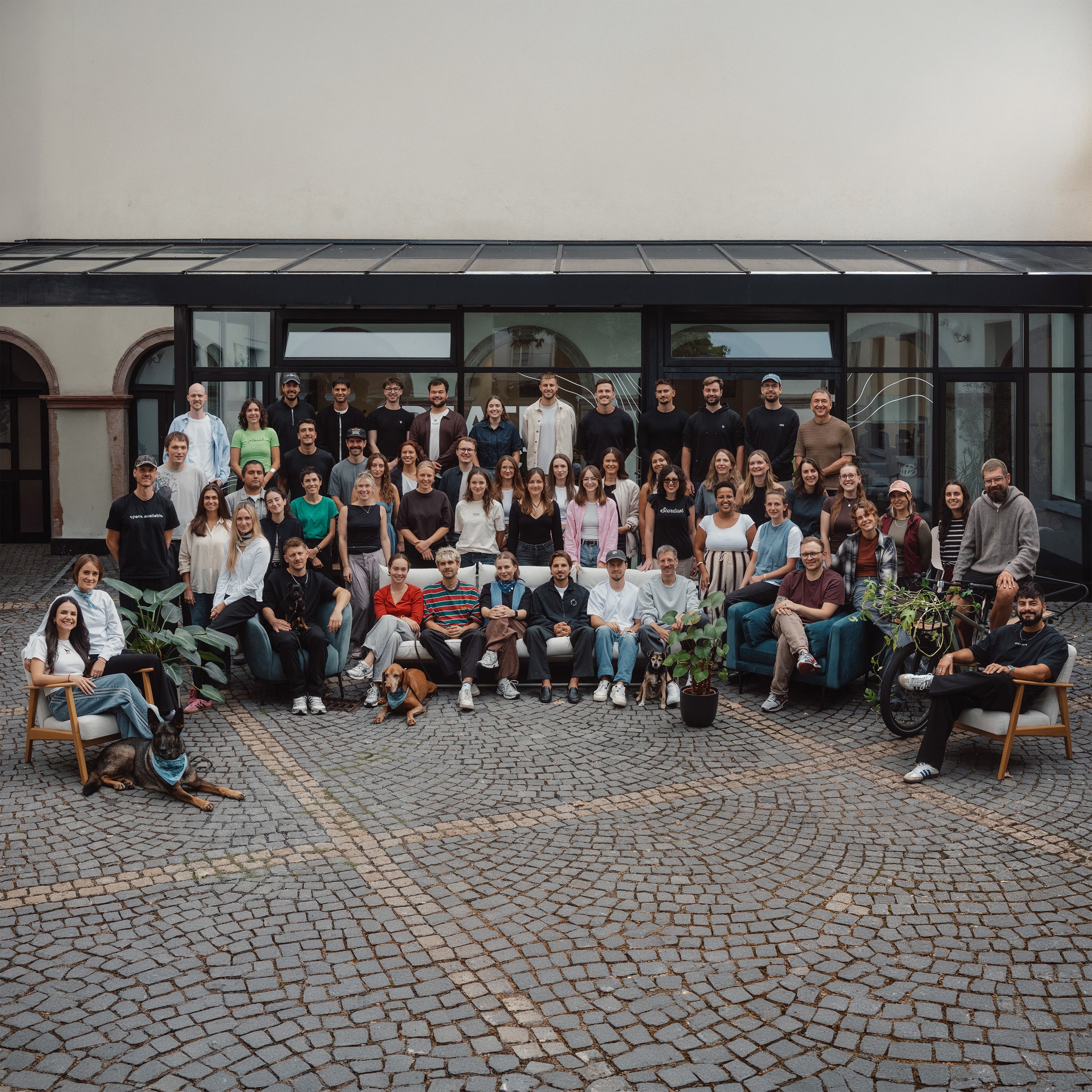 Grote teamfoto van mannen en vrouwen die buiten poseren op een geplaveide binnenplaats voor een gebouw.