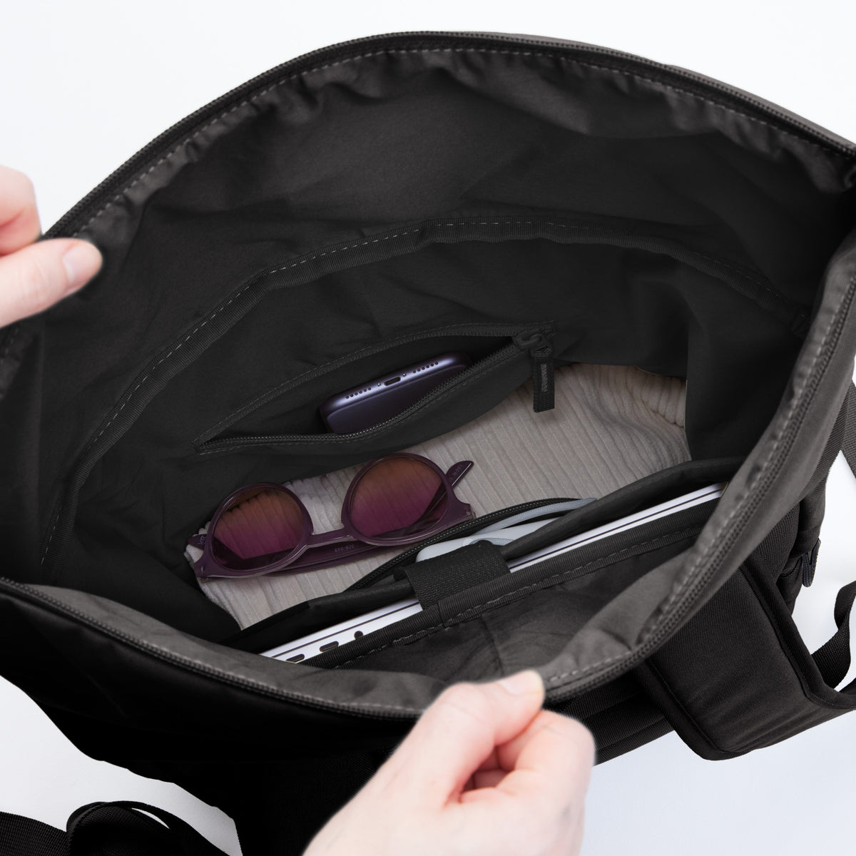 Innenansicht einer Tasche mit Smartphone, Sonnenbrille und Laptop in den Fächern, von Händen offen gehalten.