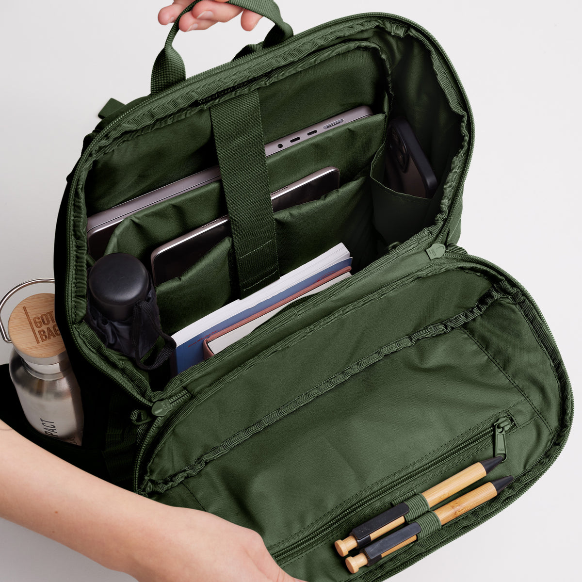 Hände halten einen Rucksack offen, der seine Innenorganisation mit Laptop, Tablet, Handy, Schirm, Flasche, Büchern und Stiften zeigt.