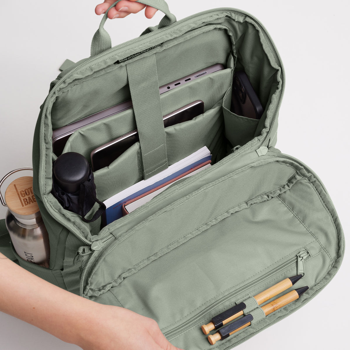 Innenansicht eines geöffneten Rucksacks, der von Händen gehalten wird, mit Laptop, Smartphone und Büchern.