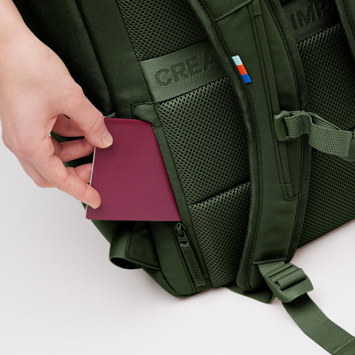 Detailaufnahme eines Rucksacks mit CREA-Logo und einer Hand, die einen bordeauxfarbenen Reisepass in ein seitliches Geheimfach steckt.